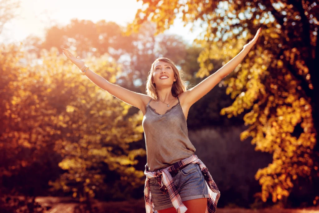 A woman standing among autumn trees with her arms raised toward the sky