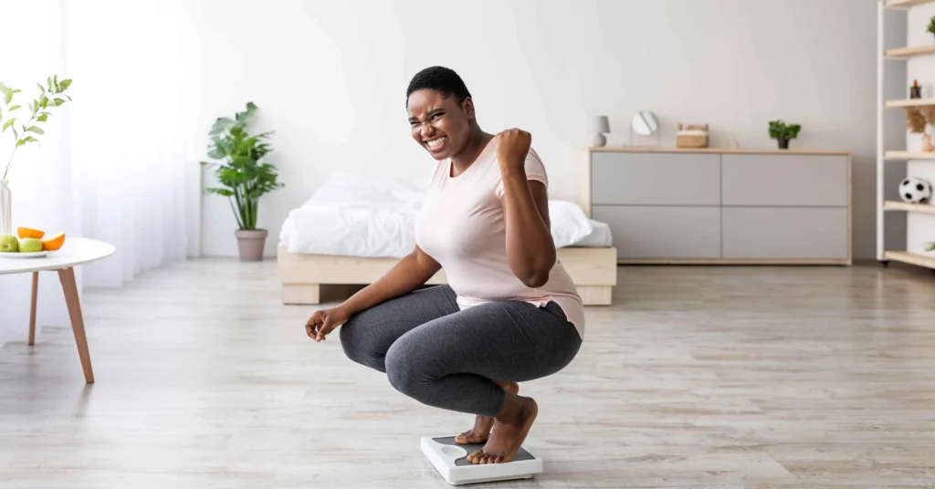 A woman crouching on a bathroom scale in a bright bedroom, celebrating